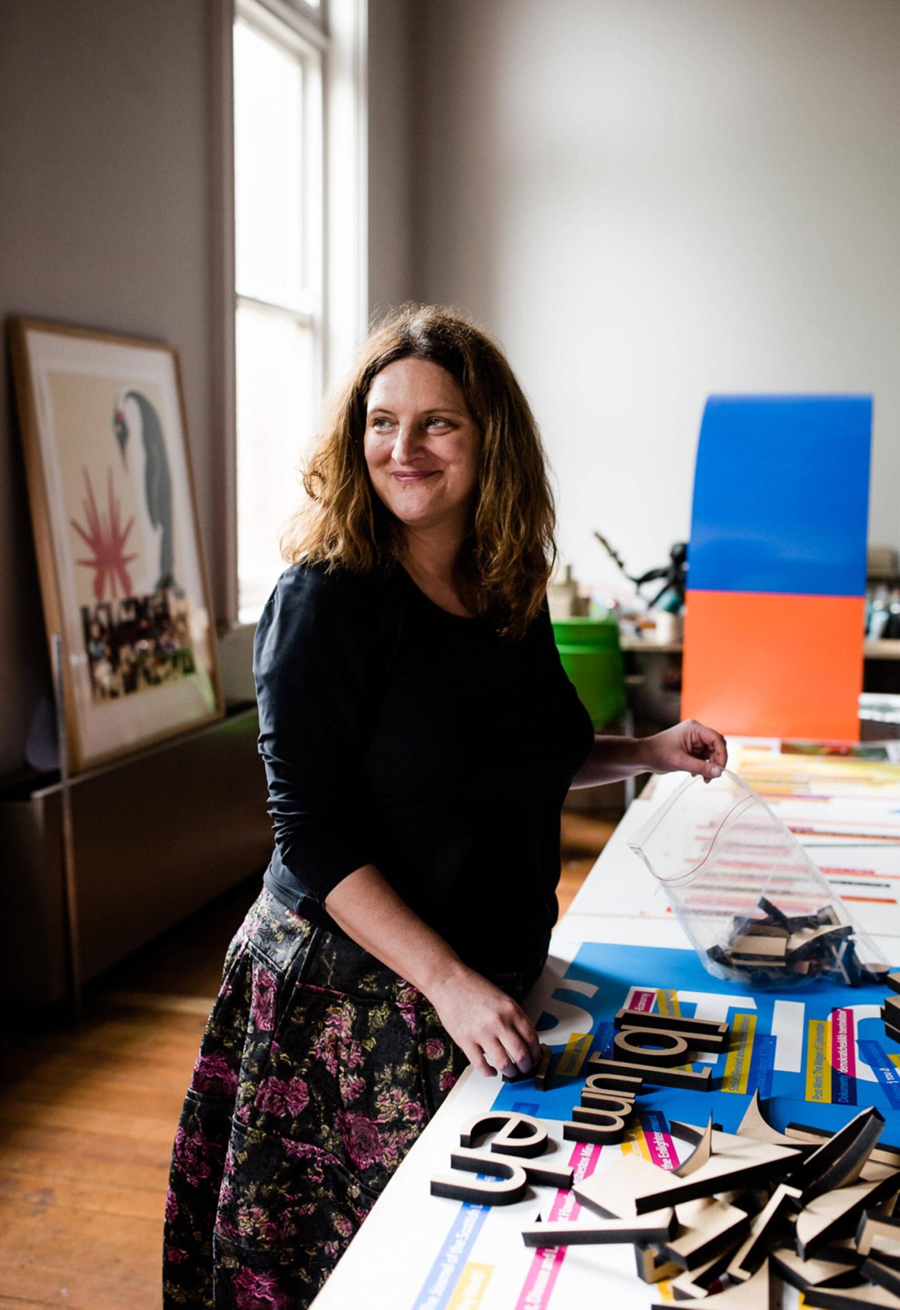 The artist stands at her worktable, smiling over her shoulder